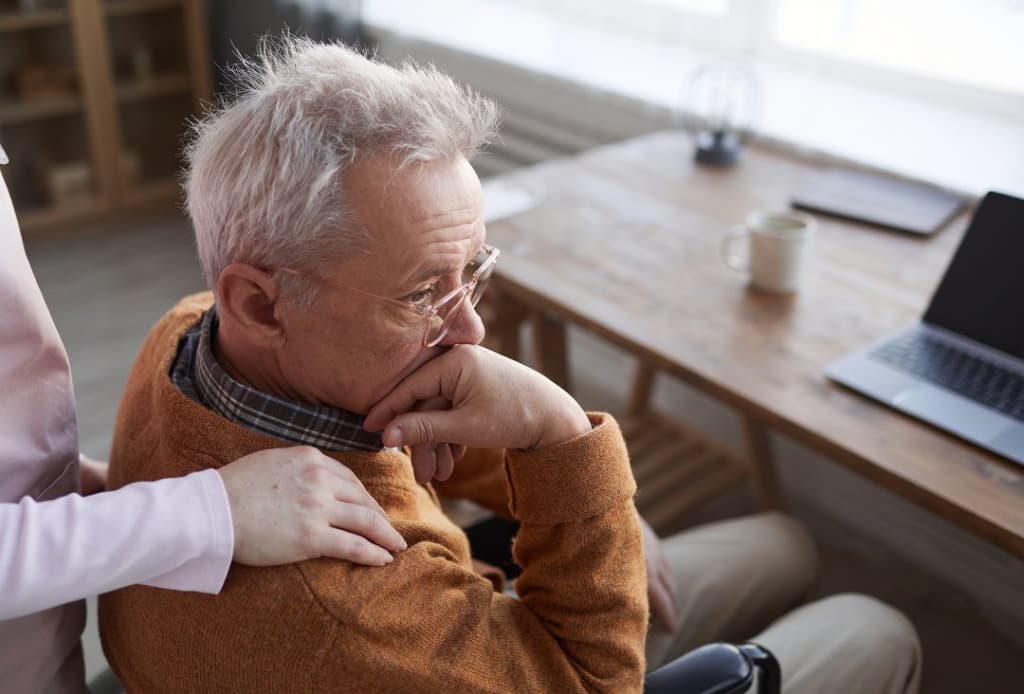 An elderly man sits looking pensive with his hand on his forehead