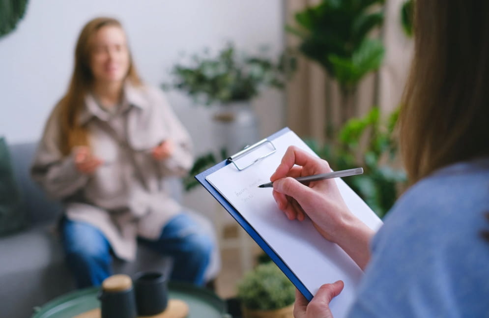 A focused therapist taking notes on a clipboard during a session with a blurred female client speaking in the background